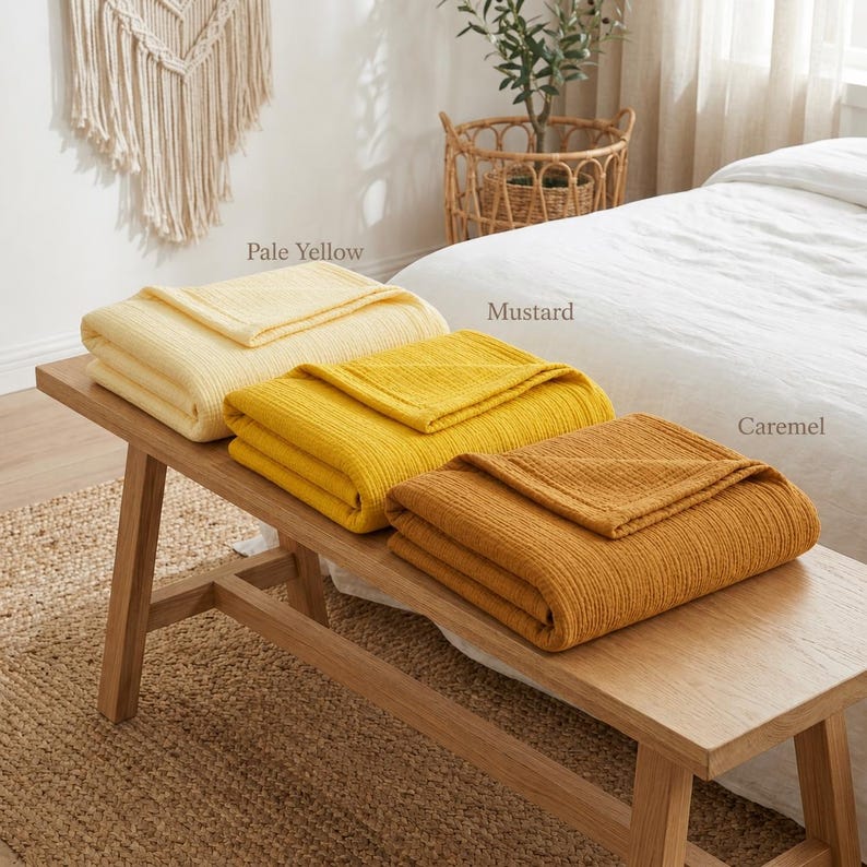 Pu&ograve; includere: Tre coperte piegate nei colori giallo pallido, senape e caramello sono esposte su una panca di legno. Le coperte hanno un design strutturato e a coste. La panca &egrave; posizionata su un tappeto tessuto, con un letto e oggetti decorativi sullo sfondo.
