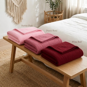 Pu&ograve; includere: Tre coperte piegate, rosa Barbie, rosa antico e rosso vino, sono esposte su una panca di legno. Le coperte hanno un aspetto strutturato. La panca &egrave; su un tappeto di iuta, con un letto e oggetti decorativi sullo sfondo.