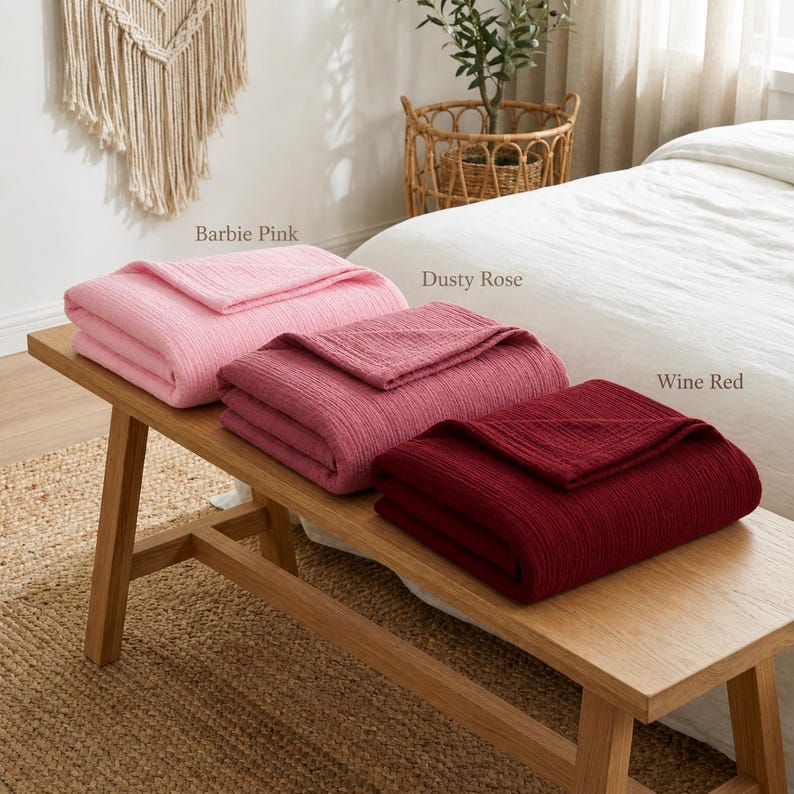 Pu&ograve; includere: Tre coperte piegate, rosa Barbie, rosa antico e rosso vino, sono esposte su una panca di legno. Le coperte hanno un aspetto strutturato. La panca &egrave; su un tappeto di iuta, con un letto e oggetti decorativi sullo sfondo.