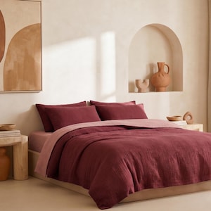 Pu&ograve; includere: Una camera da letto con un letto vestito con un piumino e cuscini bordeaux, con un lenzuolo rosa chiaro. Un comodino in legno espone un vaso e dei libri. Un dipinto astratto &egrave; appeso alla parete.
