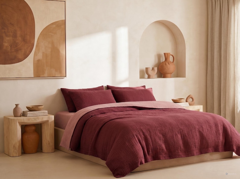 Pu&ograve; includere: Una camera da letto con un letto vestito con un piumino e cuscini bordeaux, con un lenzuolo rosa chiaro. Un comodino in legno espone un vaso e dei libri. Un dipinto astratto &egrave; appeso alla parete.