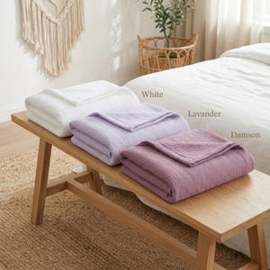 Pu&ograve; includere: Tre coperte piegate, bianche, lavanda e prugna, sono esposte su una panca di legno. Le coperte hanno un aspetto strutturato. Le parole "White", "Lavender" e "Damson" sono scritte accanto a ogni coperta. Un letto e un cesto di vimini sono sullo sfondo.