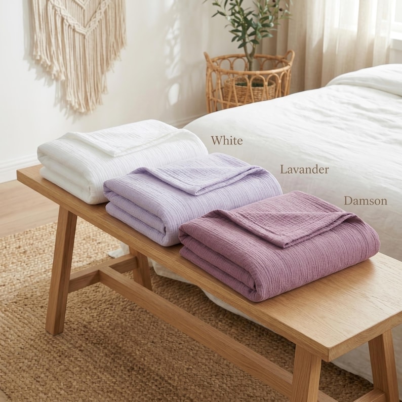 Pu&ograve; includere: Tre coperte piegate, bianche, lavanda e prugna, sono esposte su una panca di legno. Le coperte hanno un aspetto strutturato. Le parole "White", "Lavender" e "Damson" sono scritte accanto a ogni coperta. Un letto e un cesto di vimini sono sullo sfondo.