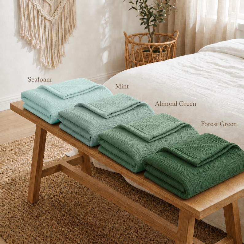 Pu&ograve; includere: Quattro coperte piegate nelle tonalit&agrave; di verde acqua, menta, verde mandorla e verde foresta sono esposte su una panca di legno. Le coperte hanno una trama strutturata e sono etichettate con i rispettivi colori. La panca &egrave; su un tappeto tessuto.