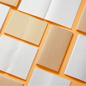 Puede incluir: Una variedad de cuadernos abiertos y cerrados con cubiertas de papel kraft y páginas blancas rayadas o cuadriculadas. Los cuadernos están dispuestos sobre un fondo naranja. Algunos cuadernos están abiertos, revelando las páginas interiores.