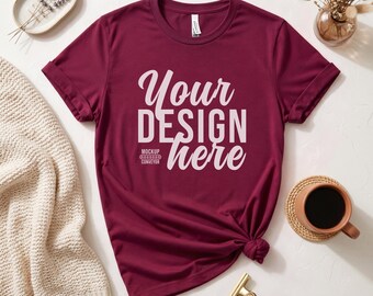 Puede incluir: Camiseta burdeos con mangas enrolladas y el texto "Your Design Here" en blanco. La camiseta está anudada en la parte inferior. Una taza de café, un clip dorado y un suéter beige también están en la imagen.