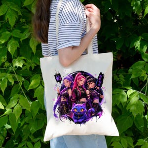 Pode incluir: Uma sacola de lona cor de creme com uma estampa gr&aacute;fica de tr&ecirc;s figuras estilizadas com cabelos roxos e rosas, acompanhadas por um pequeno gato roxo com grandes olhos amarelos. O design inclui texto em coreano e &iacute;cones de cora&ccedil;&otilde;es e estrelas.