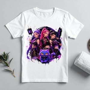 Pode incluir: Uma t-shirt branca de manga curta com uma impress&atilde;o gr&aacute;fica de tr&ecirc;s personagens estilo anime com cabelo roxo e rosa, vestidos com roupas escuras, posicionados acima de um gato roxo estilizado com grandes olhos amarelos e uma coroa.