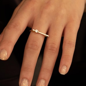 May include: A gold ring with a small, round, clear gemstone is worn on a finger. The ring is on a hand with manicured nails. The background is a dark, solid colour.