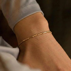 Pode incluir: Uma delicada pulseira dourada com uma série de pequenas contas cilíndricas espaçadas uniformemente e elos de conexão. A pulseira é usada no pulso, mostrando seu design minimalista e elegância sutil. A cor dourada quente contrasta com o tom de pele.