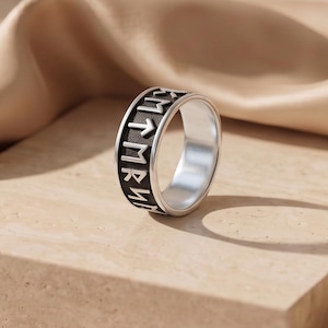 May include: A silver-toned metal ring featuring a wide band with a black recessed centre. The band is engraved with a series of distinct runic symbols in a repeating pattern around the entire circumference of the outer surface of the ring.