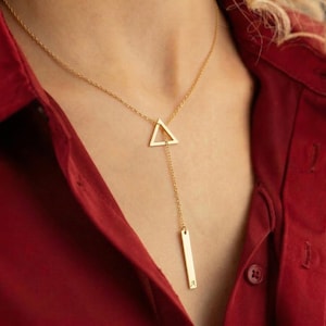 Può includere: Una collana color oro con un design geometrico. La collana presenta un ciondolo a triangolo collegato a un ciondolo a barra verticale. La collana è indossata su una camicia rossa.