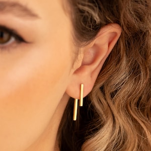 May include: Close-up of a person's ear with two gold bar earrings. The earrings are simple, linear designs, with one bar slightly longer than the other. The person has light skin and wavy, light brown hair.