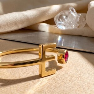 Op de afbeelding: Een goudkleurige armband met de letter "E" als designelement. De armband heeft een ronde, rode edelsteen. De achtergrond omvat een lichtgekleurde stof en een heldere kristal.