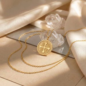 以下が含まれることがあります： 金色のペンダントネックレス。円形のペンダントには十字のデザインとテキストが施されています。ペンダントは繊細なチェーンから吊り下げられています。ネックレスは、柔らかいドレープの生地と透明なクリスタルが置かれたニュートラルな表面に展示されています。