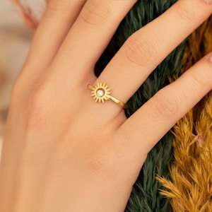 Puede incluir: Un anillo dorado con un diseño de sol y una gema clara central. El anillo se lleva en un dedo, con un fondo borroso de follaje verde y amarillo.