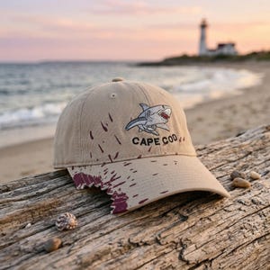 Könnte beinhalten: Beige Baseball Cap mit einem Hai-Motiv und dem Text "CAPE COD". Die Kappe hat ein Used-Design mit weinroten Farbspritzern. Der Hut liegt auf einem verwitterten Baumstamm am Strand.