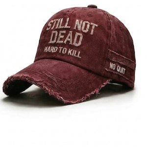 May include: A maroon baseball cap with distressed edges. The cap features the embroidered text "STILL NOT DEAD HARD TO KILL" on the front and "NO QUIT" on the side. The hat has a curved brim and a fabric strap closure.