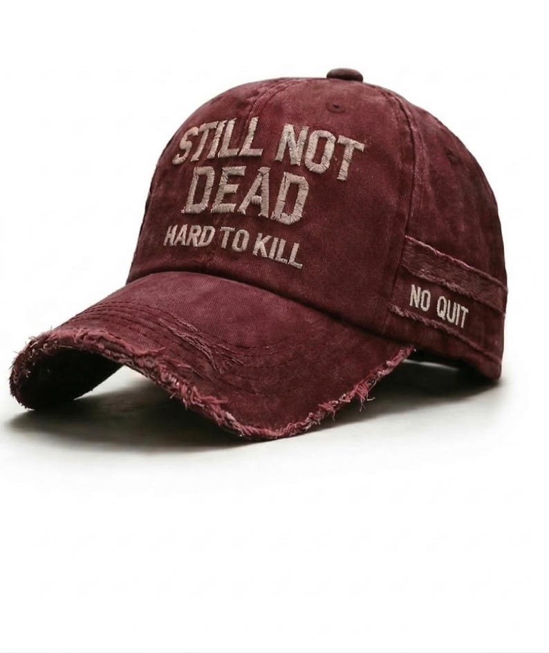 May include: A maroon baseball cap with distressed edges. The cap features the embroidered text "STILL NOT DEAD HARD TO KILL" on the front and "NO QUIT" on the side. The hat has a curved brim and a fabric strap closure.