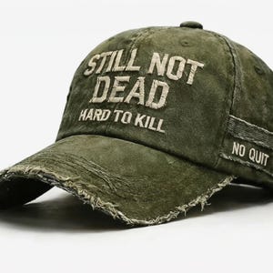 May include: Olive green baseball cap with distressed details. The cap features the embroidered text "STILL NOT DEAD HARD TO KILL" on the front and "NO QUIT" on the side. The brim has a frayed edge.