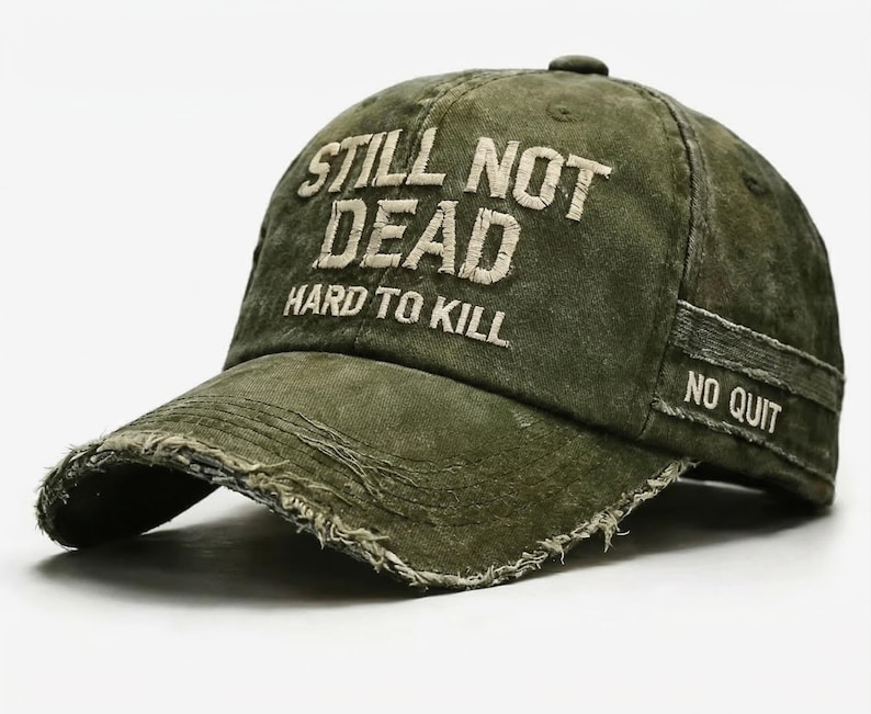 May include: Olive green baseball cap with distressed details. The cap features the embroidered text "STILL NOT DEAD HARD TO KILL" on the front and "NO QUIT" on the side. The brim has a frayed edge.