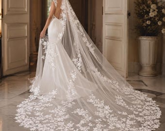May include: A floor-length white wedding dress with a fitted bodice, an open back, and a long, flowing skirt. A sheer, cathedral-length veil made of white tulle features intricate floral lace embroidery that extends along the edges and train.