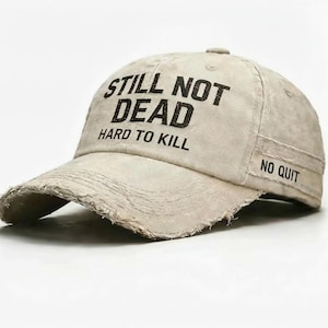May include: A beige baseball cap with distressed edges. The cap features the text "STILL NOT DEAD HARD TO KILL" across the front, with "NO QUIT" on the side. The hat has a curved brim and a slightly worn appearance.