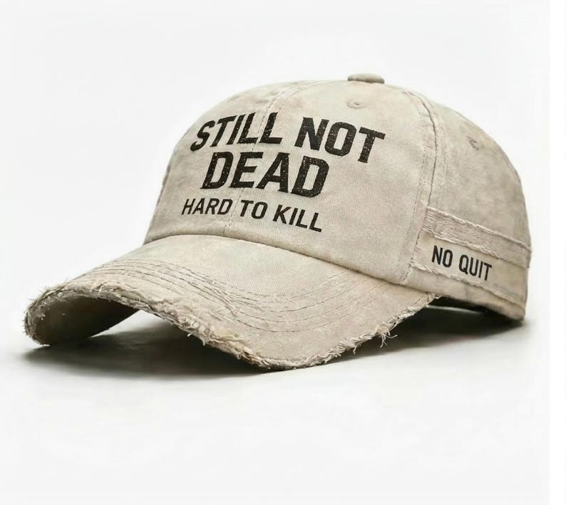 May include: A beige baseball cap with distressed edges. The cap features the text "STILL NOT DEAD HARD TO KILL" across the front, with "NO QUIT" on the side. The hat has a curved brim and a slightly worn appearance.