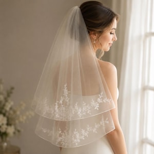 May include: A two-tier ivory tulle wedding veil featuring delicate white floral embroidery along the scalloped edges. The sheer fabric drapes softly from the crown, with the shorter top layer and longer bottom layer both adorned with intricate lace.