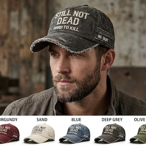 Puede incluir: Gorra de béisbol gris oscuro desgastada con el texto "STILL NOT DEAD HARD TO KILL" en blanco. La gorra tiene un borde deshilachado y las palabras "NO QUIT" en el lateral. Se muestran opciones de color adicionales.