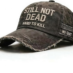 May include: A distressed, dark gray baseball cap with the text "STILL NOT DEAD HARD TO KILL" embroidered in white on the front. The brim has a frayed edge, and the side features a patch that reads "NO QUIT".