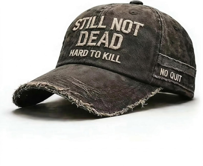 May include: A distressed, dark gray baseball cap with the text "STILL NOT DEAD HARD TO KILL" embroidered in white on the front. The brim has a frayed edge, and the side features a patch that reads "NO QUIT".