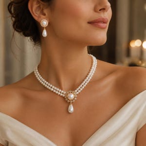 Può includere: Una collana di perle a doppio filo con un ciondolo circolare centrale caratterizzato da un gruppo di piccole pietre trasparenti attorno a una singola perla e una perla a goccia pendente. Gli orecchini abbinati presentano una piccola perla.