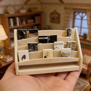 Może przedstawiać: Miniaturowy drewniany regał na książki wypełniony maleńkimi książkami. Regał ma trzy poziomy i mieści kilka małych książek o różnych wzorach okładek. Książki są ułożone na półkach, tworząc uroczy wystrój. Scena rozgrywa się w domku dla lalek.