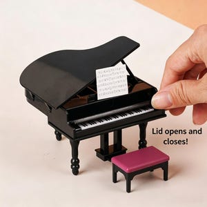Pode incluir: Um piano de cauda em miniatura preto com teclado branco, uma estante com partitura e um pequeno banco separado com assento magenta. O piano possui uma tampa articulada que abre e fecha, conforme indicado pelo texto Lid opens and closes!