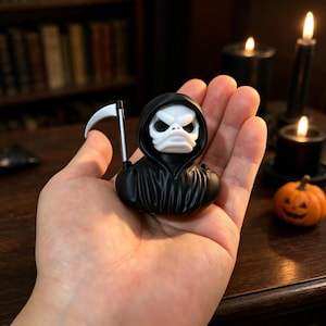 Puede incluir: Una pequeña figura de pato de goma con diseño de parca, que presenta una capa negra con capucha, una cara blanca similar a una calavera con expresión de enfado y una guadaña de color plateado unida al lateral. Es compacta y de plástico liso.