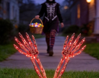 Puede incluir: Dos manos de esqueleto iluminadas que emergen de la hierba, con luces rojas brillantes dentro de la estructura ósea de plástico. Las manos están colocadas en posición vertical con los dedos extendidos, creando un efecto brillante y siniestro.