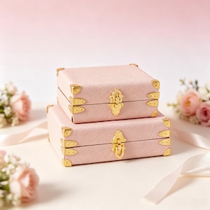 May include: Two rectangular light pink storage boxes with a textured finish, stacked one on top of the other. Each box features gold-toned metal corner protectors and a central gold-toned metal latch mechanism on the front panel.