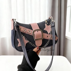 Puede incluir: Bolso de hombro negro y rosa con detalles de estrellas y cierre de hebilla. El bolso presenta una correa rosa y un detalle de cadena plateada. El cuerpo del bolso es azul oscuro con adornos de estrellas rosas.