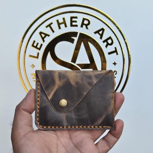 Könnte beinhalten: Ein brauner Kartenhalter aus Leder mit einer abgenutzten Textur, der eine dreieckige Klappe mit einem einzelnen messingfarbenen Metalldruckknopf besitzt. Die Kanten sind mit dickem, hellem Garn vernäht, was einen sichtbaren Rand erzeugt.
