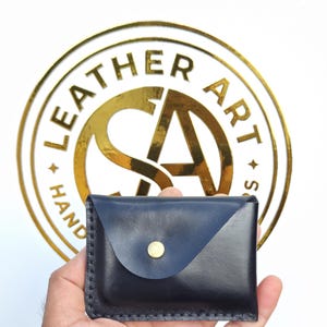 May include: A dark navy blue leather card holder with a triangular flap closure secured by a single brass-colored metal snap button. The edges are stitched with dark thread, and the leather has a smooth, polished finish with a visible sheen.