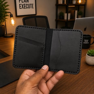 May include: A black leather bifold wallet featuring visible saddle stitching along the edges, two interior card slots, and a central compartment. The wallet has a smooth matte finish and rounded corners, showcasing a minimalist construction.