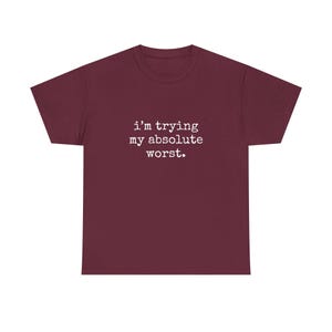 May include: A maroon t-shirt featuring the white text "i'm trying my absolute worst". The text is centered on the chest. The shirt has a crew neck and short sleeves.