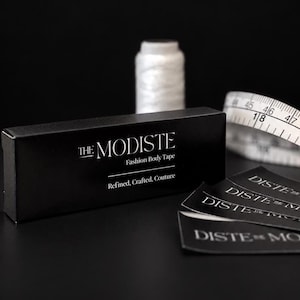 May include: A black rectangular box labelled THE MODISTE Fashion Body Tape with the text Refined, Crafted, Couture. Beside the box are several black strips with white text and a white measuring tape showing 18 inches and 48 cm markings.