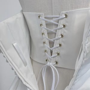 May include: A white corset-style bodice featuring a lace-up back closure with silver-toned metal grommets and white ribbon lacing. The garment includes a structured panel with vertical stitching and a row of fabric-covered buttons along the side.