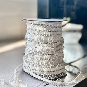May include: A spool of white button loop trim featuring a continuous series of small, uniform fabric loops attached to a flat woven tape. A metal ruler is visible at the base, showing measurements in centimetres, with the trim extending outward.