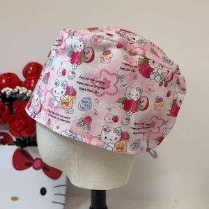 Op de afbeelding: Een roze operatiemuts met een herhaald patroon van Hello Kitty-personages, aardbeien, cupcakes en melkpakken. De muts heeft de tekst "Around the galaxy, there are no brighter stars than you." en "I hope you're here for me."