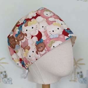 Op de afbeelding: Een patroon scrub cap met een herhalend ontwerp van cartoonberen en katten in roze, rood en blauw. De cap wordt getoond op een mannequinhoofd met een houten standaard.
