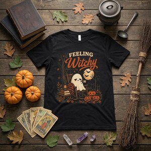 Può includere: Una t-shirt nera con il testo "FEELING Witchy" sopra un'illustrazione di un fantasma seduto su una sedia a dondolo, circondato da una lanterna, libri, zucche, una luna piena con pipistrelli e alberi nei toni dell'arancione e del marrone.