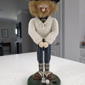 May include: A wooden nutcracker figurine dressed as a golfer, wearing a dark blue beret, a cream-colored knit sweater, dark blue trousers, and argyle socks. The figure holds a miniature golf club over a green base with a small golf ball.